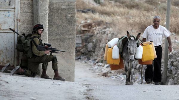 Viro transportas akvon azendorse, antaŭ soldatoj de la okupacia armeo en operaco, en vilaĝo de Cisjordanio - © AFP