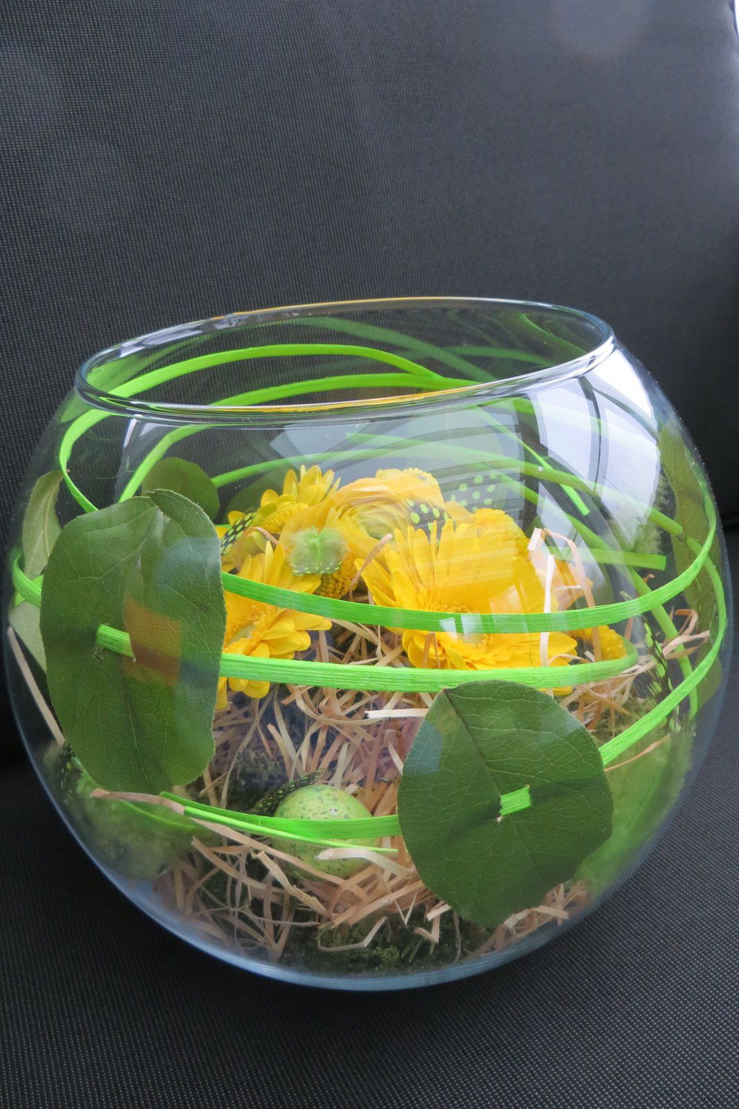 Base en verre, mousse, paille ,rotin, plumes et végétaux : renoncules, gerbera , santini et feuilles enfilées sur le rotin.